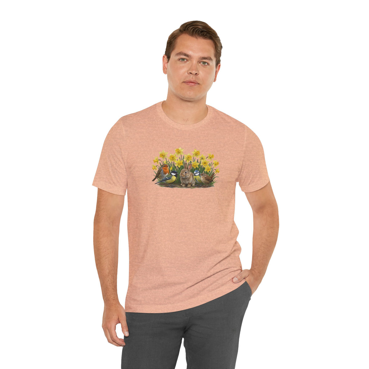 Mann trägt ein peach T-Shirt mit buntem Vogel- und Kaninchen-Motiv in einer Frühlingsumgebung.