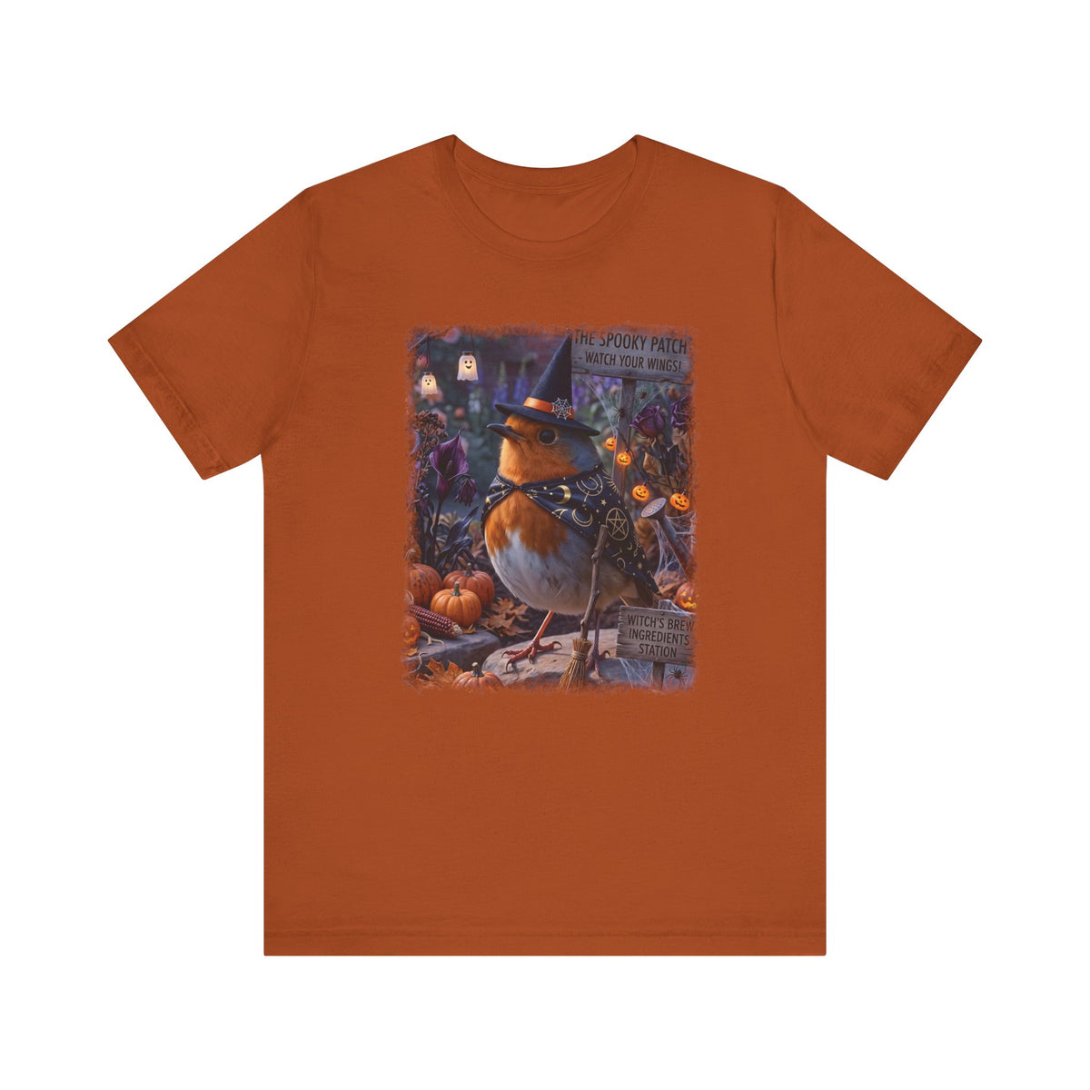 An orange short-sleeve crewneck t-shirt featuring a central graphic print of a robin bird dressed as a witch sitting among pumpkins and autumn foliage.