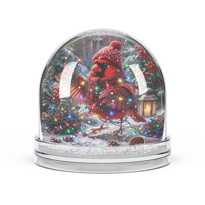 A clear glass snow globe with a silver base, featuring a painted red cardinal wearing a knit hat and scarf, surrounded by Christmas lights, pine trees, berries, and a lantern inside.