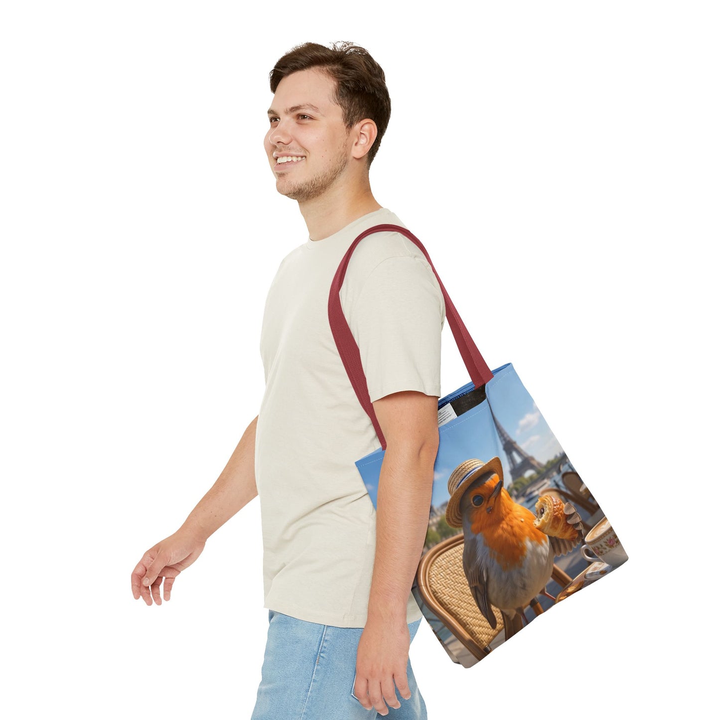 A man in a light beige t-shirt and blue jeans is shown from the waist up, carrying a canvas tote bag over his shoulder. The tote bag features a printed design of a robin wearing a straw hat on a wicker basket, with a blue sky background and a dark red strap.