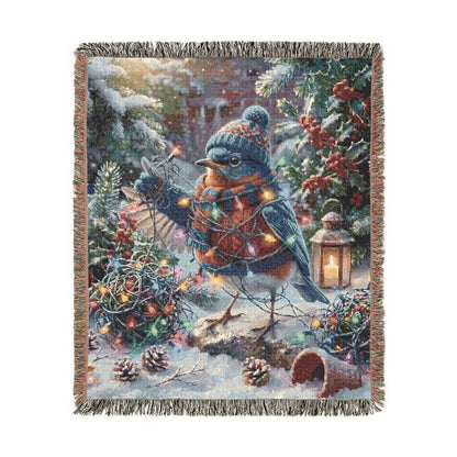 A rectangular woven throw blanket with fringed edges, featuring a whimsical winter forest scene with a bluebird, evergreen branches, pinecones, red berries, and warm string lights, shown on a plain white background.