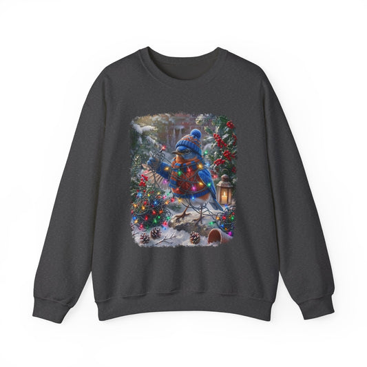 A dark gray crewneck sweatshirt featuring a large square graphic print on the front. The graphic depicts a festive winter scene with a bluebird wearing a blue knit hat and scarf, surrounded by decorated Christmas trees, pinecones, and a lantern in the snow.