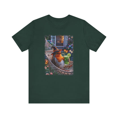 A dark forest green short-sleeve crewneck t-shirt featuring a colorful graphic print of a robin bird relaxing in a hammock surrounded by Halloween decorations like jack-o'-lanterns and a green drink.