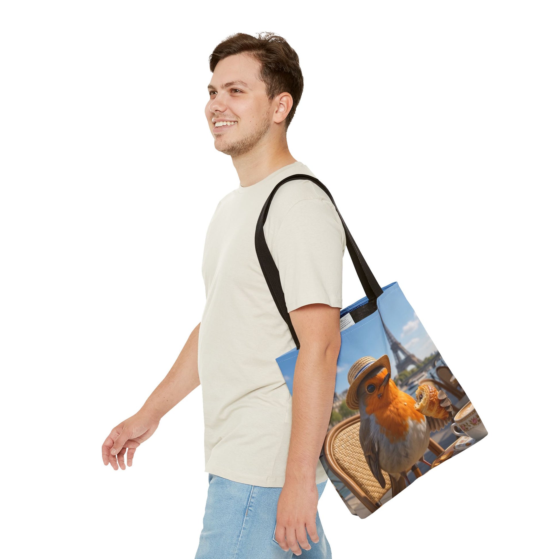 A man in a light beige t-shirt and blue jeans is shown from the waist up, carrying a canvas tote bag over his shoulder. The tote bag features a colorful printed design of a robin wearing a hat in front of the Eiffel Tower.