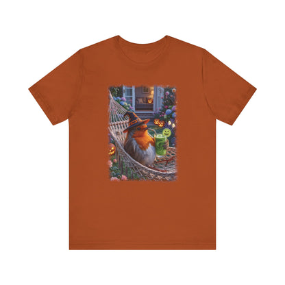 An orange short-sleeve crewneck t-shirt featuring a central graphic print of a robin bird wearing a witch hat sitting in a hammock surrounded by Halloween decorations.