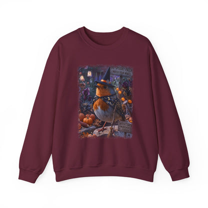 A maroon crewneck sweatshirt featuring a colorful graphic print of a robin bird wearing a witch hat on a pumpkin stump in front of a spooky house, shown against a plain white background.
