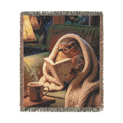 A woven throw blanket with fringed edges, featuring a cozy scene of a robin bird wrapped in a beige knit blanket, reading a book and holding a mug. The blanket is photographed flat against a plain white background.