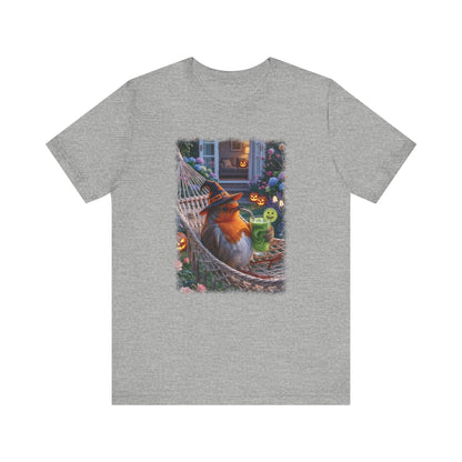 A heather gray short-sleeve crewneck t-shirt featuring a large rectangular graphic print on the front. The graphic depicts a cozy Halloween scene with a fluffy robin bird wearing a witch hat, lounging in a hammock next to a green glowing drink.
