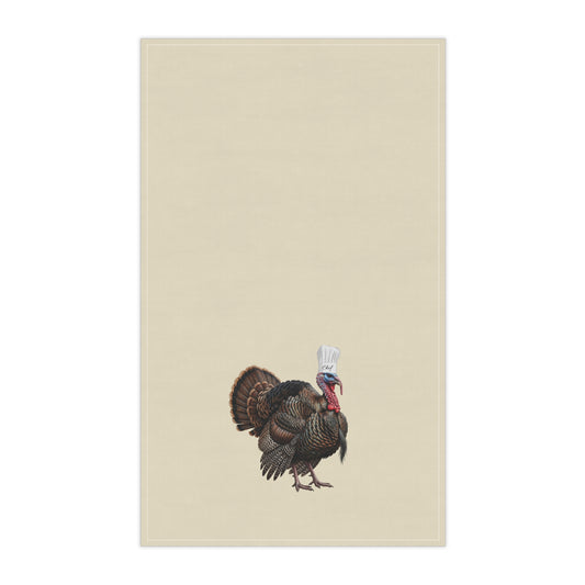 A rectangular beige kitchen towel featuring a small illustrated turkey near the bottom center, wearing a white chef's hat. The product is shown flat against a plain white background.