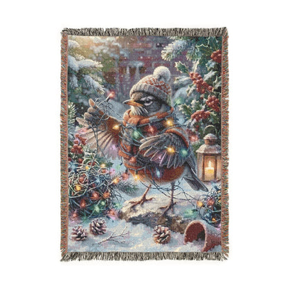 A rectangular woven throw blanket with fringed edges, featuring a whimsical winter woodland scene with a robin in a hat and scarf, pinecones, berries, evergreen branches, and warm string lights on a white background.