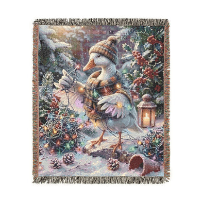A square woven throw blanket with fringed edges, featuring a whimsical winter scene with a goose in a striped sweater and scarf, surrounded by snow, evergreen branches, pinecones, and warm string lights.
