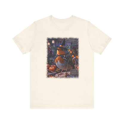 An off-white crewneck t-shirt featuring a colorful graphic print of a robin dressed as a witch surrounded by pumpkins on a white background.