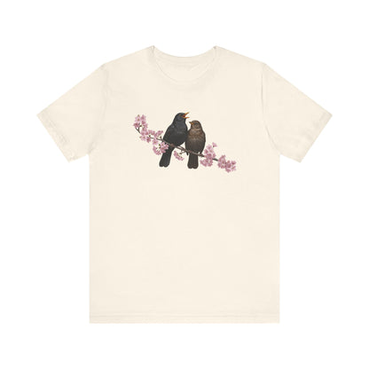 Beige t-shirt with a graphic of two birds on a branch with pink flowers on a white background
