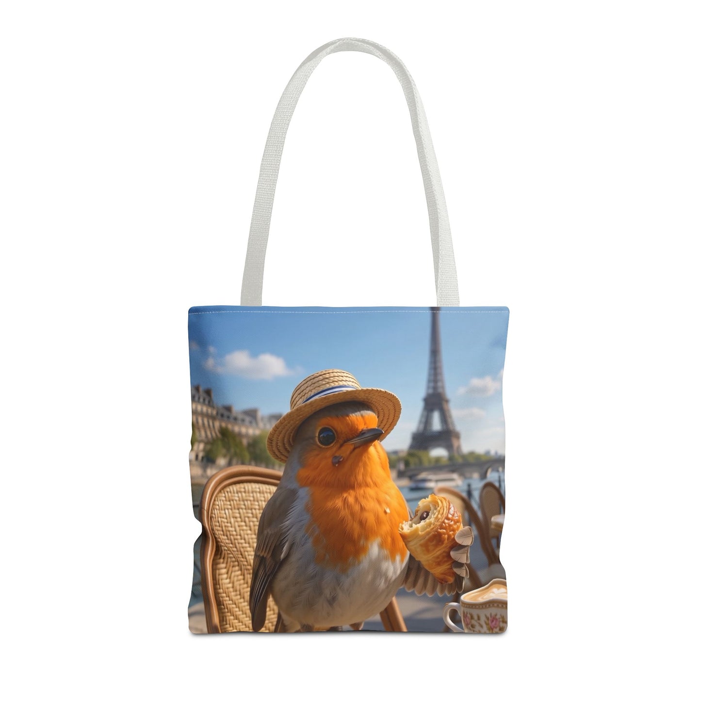 A white canvas tote bag featuring a printed graphic of a robin wearing a straw hat and holding a croissant, with the Eiffel Tower and Parisian cafe tables in the background.