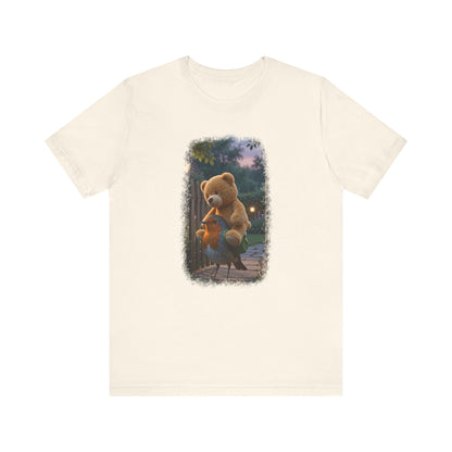 Naturfarbenes T-Shirt aus Baumwolle. Auf der Brust ist eine witzige digitale Illustration aufgedruckt: Ein niedliches Rotkehlchen namens Rosi steht im Vorgarten und versucht, einen riesigen Kirmes-Teddybären in einen viel zu kleinen grünen Frosch-Rucksack zu quetschen.