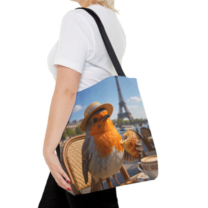 A woman wearing a white t-shirt and black pants carries a canvas tote bag over her shoulder. The tote bag features a printed graphic of a robin wearing a straw hat and holding a croissant, with the Eiffel Tower and a cafe table in the background.