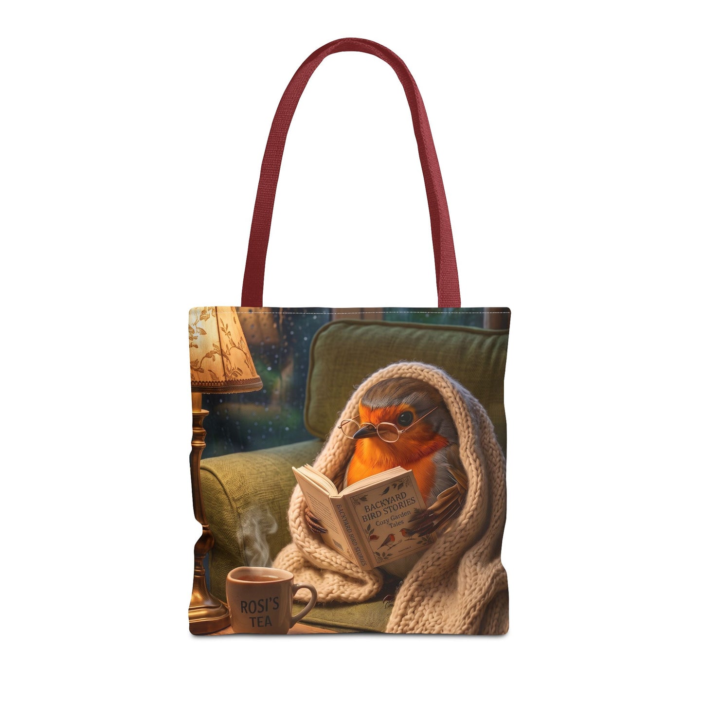 A tote bag with dark red handles, featuring a printed illustration of a robin bird wearing glasses and reading a book wrapped in a knitted blanket on a green armchair, with a steaming cup nearby.