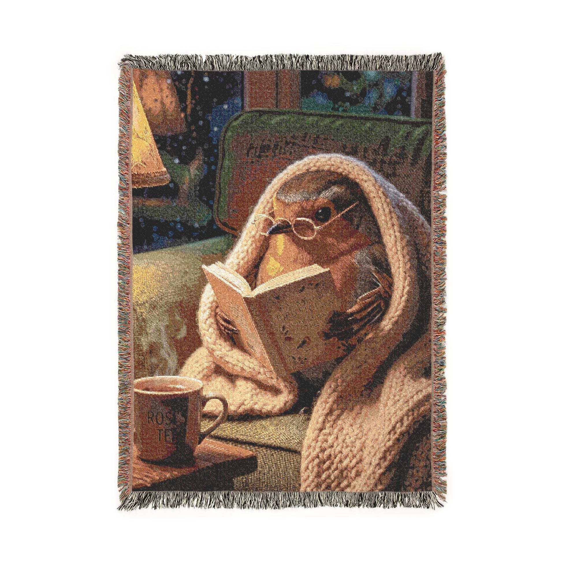 A rectangular woven throw blanket with fringed edges, featuring a cozy illustrated scene of a robin bird wrapped in a knitted blanket and scarf, reading a book with a mug nearby. The blanket is shown against a plain white background.