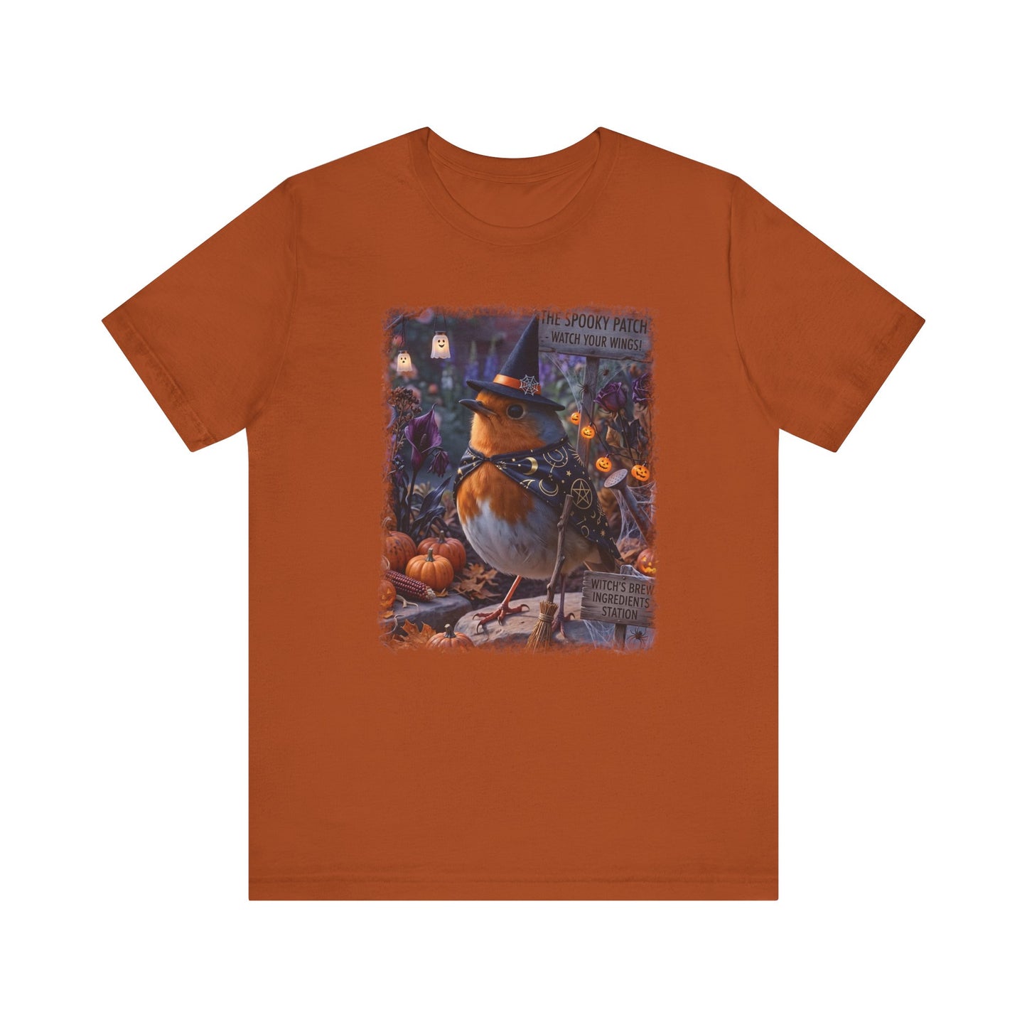 An orange short-sleeve crewneck t-shirt featuring a central graphic print of a robin bird dressed as a witch sitting among pumpkins and autumn foliage.
