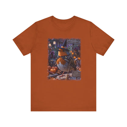 An orange short-sleeve crewneck t-shirt featuring a central graphic print of a robin bird dressed as a witch sitting among pumpkins and autumn foliage.