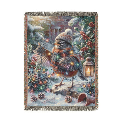 A rectangular woven throw blanket with fringed edges, featuring a whimsical winter scene with a robin, evergreen branches, pinecones, and warm string lights on a white background.