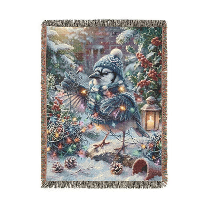 A rectangular woven throw blanket with fringed edges, featuring a winter woodland scene with a blue jay, pinecones, holly, evergreen branches, and warm string lights on a white background.