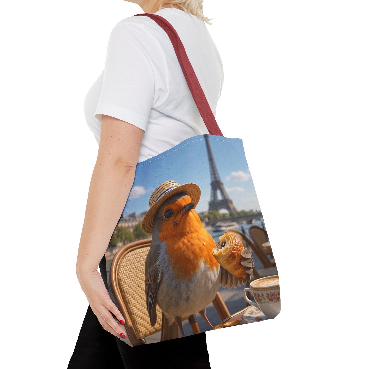 A woman wearing a white t-shirt and black pants carries a canvas tote bag over her shoulder. The bag features a printed graphic of a robin wearing a straw hat and holding a croissant, with the Eiffel Tower and a Parisian cafe table in the background.