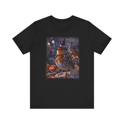 A black short-sleeve crewneck t-shirt featuring a large, colorful graphic print on the front. The graphic depicts a robin wearing a witch's hat in a spooky, autumnal garden scene with pumpkins and signs.