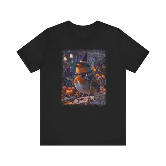 A black short-sleeve crewneck t-shirt featuring a large, colorful graphic print on the front. The graphic depicts a robin wearing a witch's hat in a spooky, autumnal garden scene with pumpkins and signs.