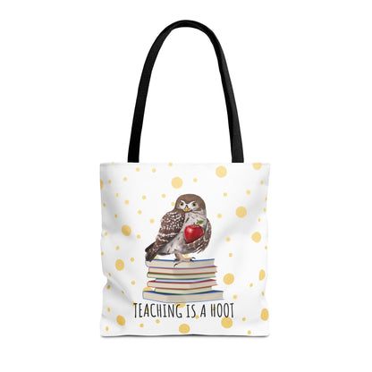 A white tote bag with black shoulder straps, featuring a printed design of a small owl perched on a stack of books and holding a red heart, with the text 'TEACHING IS A HOOT' below. The bag is decorated with scattered gold polka dots on a white background.