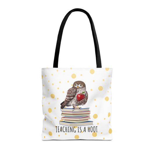 A white tote bag with black shoulder straps, featuring a printed design of a small owl perched on a stack of books and holding a red heart, with the text 'TEACHING IS A HOOT' below. The bag is decorated with scattered gold polka dots on a white background.
