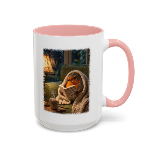 A white ceramic mug with a pink interior and handle, featuring a printed illustration of a cozy robin wrapped in a blanket and reading a newspaper on a green armchair.