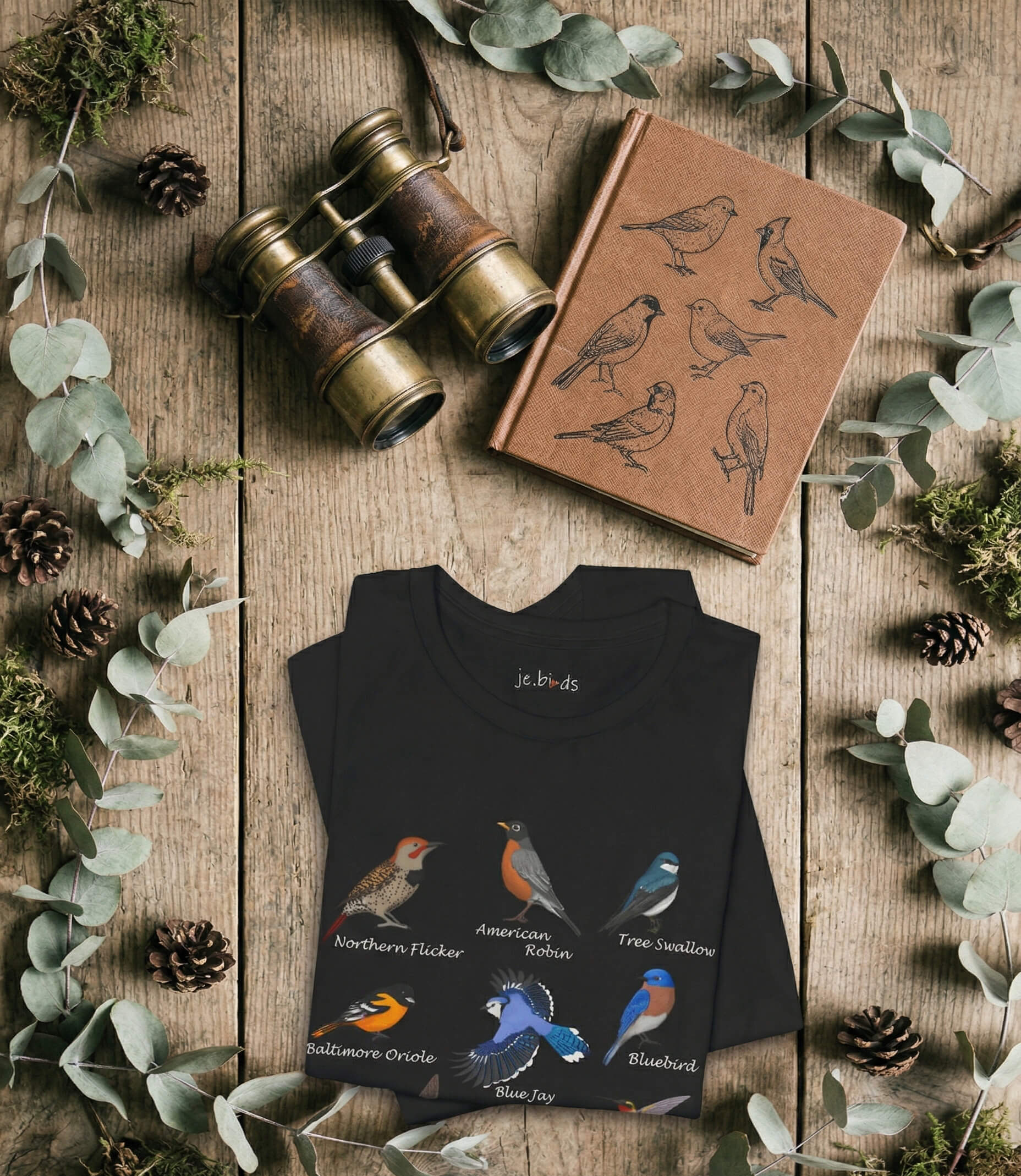 Flat lay showing The Field Guide Series t-shirt by jz.birds with vintage backyard bird illustrations like the Robin and Blue Jay, arranged with old binoculars, a leather notebook, leaves, and moss.